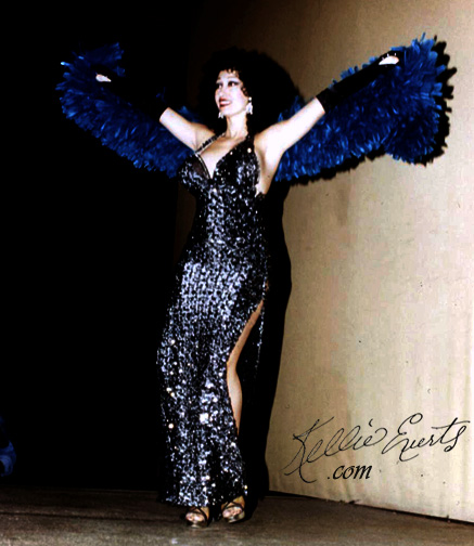 Kellie Evers the Stripper for God. Stripper / Burlesque star on stage with arms wrapped in blue frilly boa, spread like an eagle in shimmering silver gown with slit , on stage. 